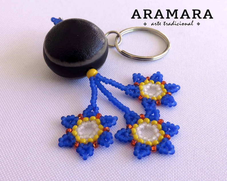 K&ouml;nnte beinhalten: Ein Schl&uuml;sselanh&auml;nger aus schwarzem und wei&szlig;em Stein mit drei daran befestigten Blumen aus blauen, gelben und orangefarbenen Perlen. Der Schl&uuml;sselanh&auml;nger ist mit "ARAMARA arte tradicional" beschriftet.