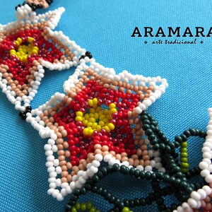 May include: Close-up of a beaded necklace with red, white, yellow, and green beads. The beads are arranged in a flower pattern. The necklace is labeled "ARAMARA arte tradicional".
