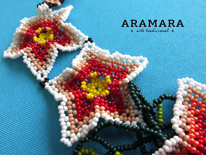 May include: Close-up of a beaded necklace with red, white, yellow, and green beads. The beads are arranged in a flower pattern. The necklace is labeled "ARAMARA arte tradicional".