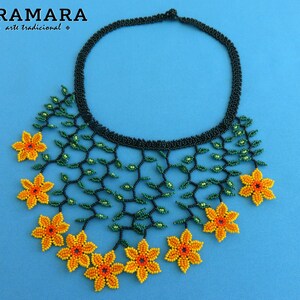 Op de afbeelding: Een kralenketting met een zwarte kraag en een bloemenontwerp. De ketting heeft groene kralenranken en geel-oranje kralenbloemen. De merknaam "ARAMARA arte tradicional" staat linksboven.