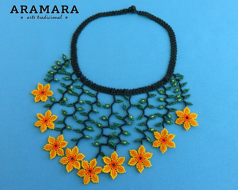 Op de afbeelding: Een kralenketting met een zwarte kraag en een bloemenontwerp. De ketting heeft groene kralenranken en geel-oranje kralenbloemen. De merknaam "ARAMARA arte tradicional" staat linksboven.