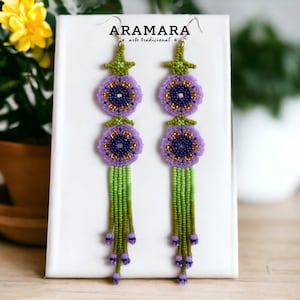 Op de afbeelding: Een paar lange, geperde oorbellen met een bloemmotief. De oorbellen hebben twee paarse bloemen met gouden centra en groene bladeren, gevolgd door een lange groene kwast. De oorbellen worden getoond op een witte kaart met de tekst "ARAMARA arte tradicional".