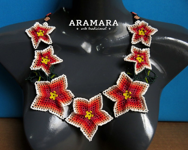 May include: A beaded necklace with a floral design. The necklace is made of small, colorful beads and features a series of red, orange, and white flowers. The flowers are arranged in a circular pattern around the neck. The necklace is on a gray mannequin.