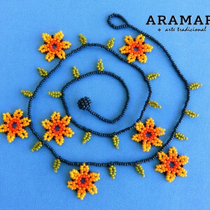 May include: A black beaded necklace with orange and yellow flower-shaped beads and green leaf-shaped beads. The necklace is coiled in a spiral shape. The text "ARAMARA * arte tradicional *" is visible in the top right corner of the image.