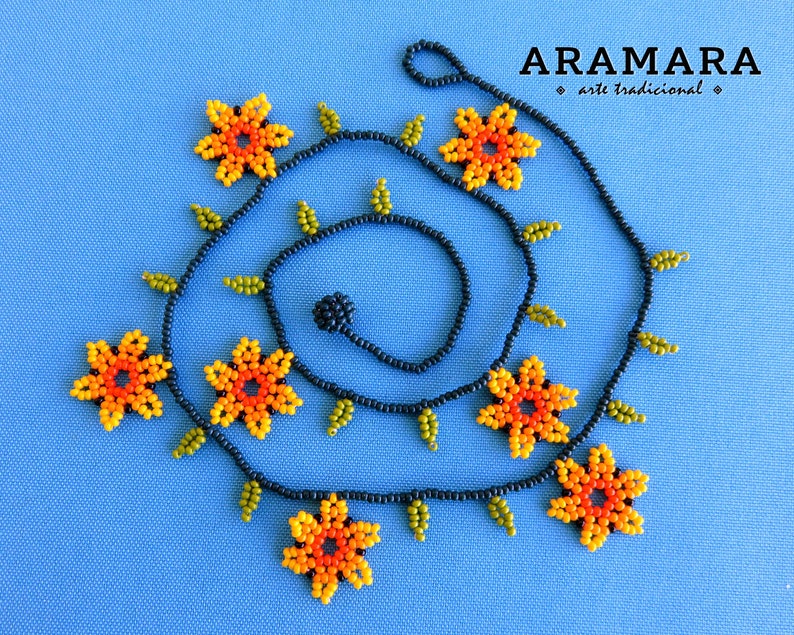 May include: A black beaded necklace with orange and yellow flower-shaped beads and green leaf-shaped beads. The necklace is coiled in a spiral shape. The text "ARAMARA * arte tradicional *" is visible in the top right corner of the image.