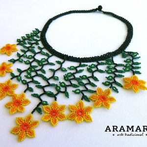 Op de afbeelding: Een kralenketting met een donkergroene kraag en een bloemmotief. De ketting heeft groene kralenranken en geel-oranje kralenbloemen. De tekst "ARAMARA arte tradicional" staat onderaan.