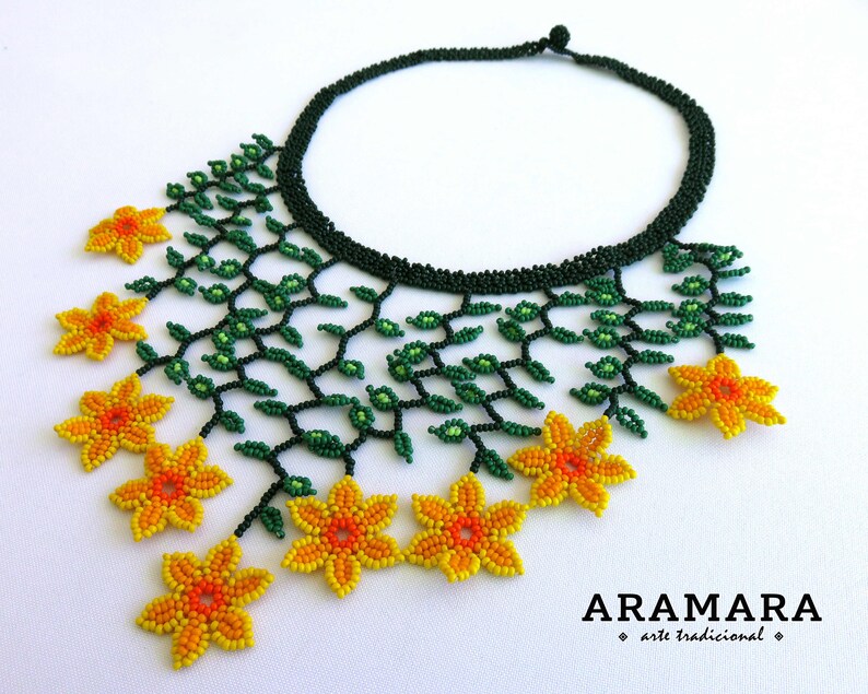 Op de afbeelding: Een kralenketting met een donkergroene kraag en een bloemmotief. De ketting heeft groene kralenranken en geel-oranje kralenbloemen. De tekst "ARAMARA arte tradicional" staat onderaan.