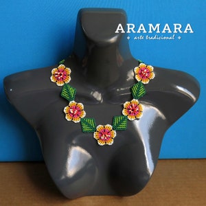 May include: A beaded necklace with a floral design. The necklace is made with pink, yellow, white, and green beads. The necklace is on a gray mannequin bust. The text "ARAMARA arte tradicional" is visible in the top right corner of the image.