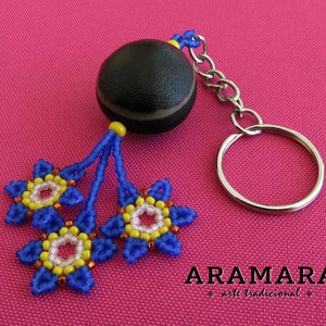 K&ouml;nnte beinhalten: Ein blauer und gelber Perlen-Schl&uuml;sselanh&auml;nger mit einem schwarzen und braunen Samenkapsel-Anh&auml;nger. Der Schl&uuml;sselanh&auml;nger hat einen silbernen Schl&uuml;sselring und der Text "ARAMARA arte tradicional" ist unter dem Anh&auml;nger gedruckt.