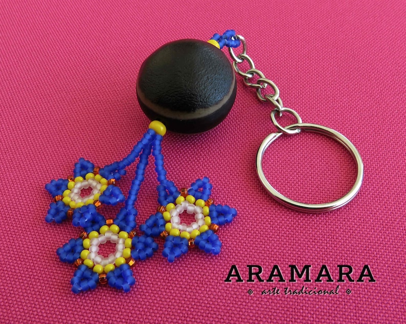 K&ouml;nnte beinhalten: Ein blauer und gelber Perlen-Schl&uuml;sselanh&auml;nger mit einem schwarzen und braunen Samenkapsel-Anh&auml;nger. Der Schl&uuml;sselanh&auml;nger hat einen silbernen Schl&uuml;sselring und der Text "ARAMARA arte tradicional" ist unter dem Anh&auml;nger gedruckt.
