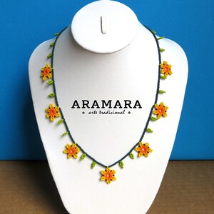 May include: A beaded necklace with yellow and orange flowers and green leaves on a dark blue string. The necklace is displayed on a white mannequin bust. The text "ARAMARA arte tradicional" is printed on the bust.