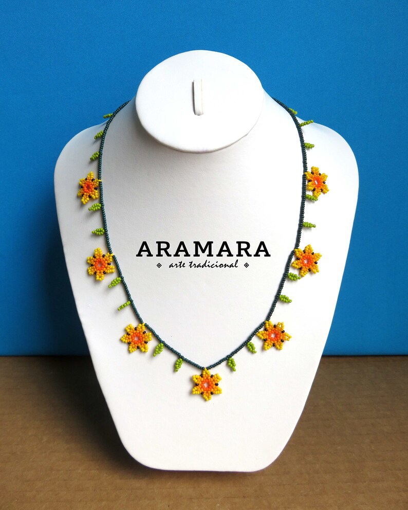 May include: A beaded necklace with yellow and orange flowers and green leaves on a dark blue string. The necklace is displayed on a white mannequin bust. The text "ARAMARA arte tradicional" is printed on the bust.
