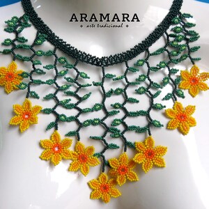 Op de afbeelding: Een kralenketting met een bloemenmotief. De ketting heeft een donkergroene kralenbasis met groene kralenranken en geel-oranje kralenbloemen. De tekst "ARAMARA arte tradicional" staat linksboven.