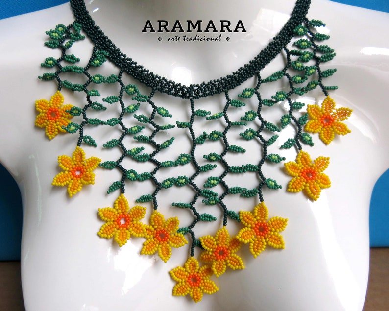Op de afbeelding: Een kralenketting met een bloemenmotief. De ketting heeft een donkergroene kralenbasis met groene kralenranken en geel-oranje kralenbloemen. De tekst "ARAMARA arte tradicional" staat linksboven.