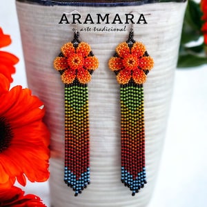 Puede incluir: Un par de aretes largos de cuentas con un diseño de flores. Las flores son de color naranja, rojo y amarillo, y las cuentas son de color negro, verde, naranja, rojo, amarillo, azul y turquesa. Los aretes están etiquetados como "ARAMARA - arte tradicional".