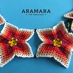 May include: A close-up of a beaded necklace with a star-shaped flower design. The necklace is made of red, orange, yellow, and white beads. The text "ARAMARA arte tradicional" is visible in the background.