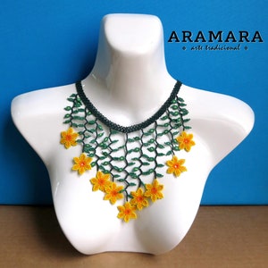 Op de afbeelding: Een kralenketting op een witte mannequin. De ketting heeft een donkergroene kralen kraag, groene ranken en felgele bloemaccenten. De tekst "ARAMARA arte tradicional" is zichtbaar in de afbeelding.
