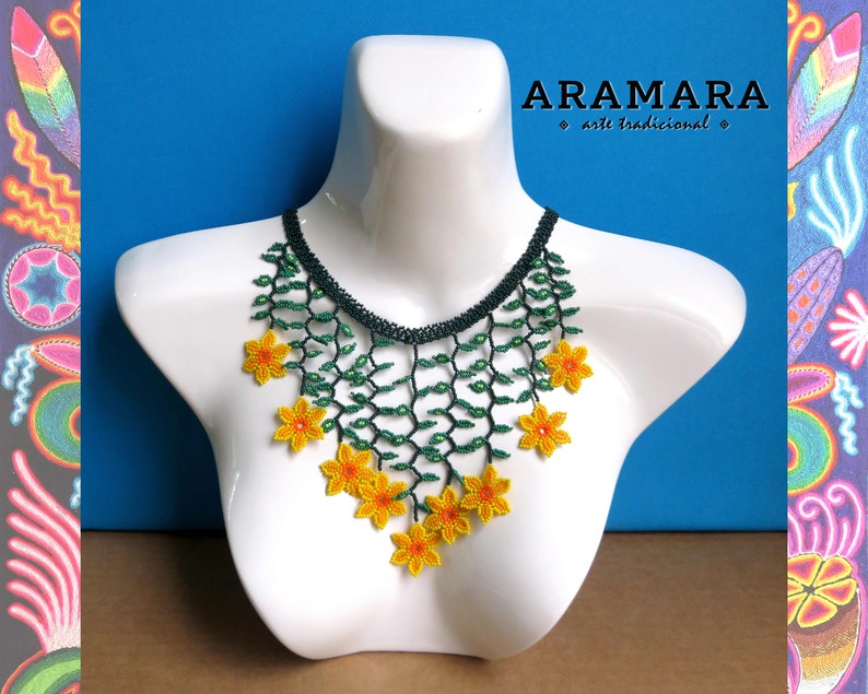 Op de afbeelding: Een kralenketting op een witte mannequin. De ketting heeft een donkergroene kralen kraag, groene ranken en felgele bloemaccenten. De tekst "ARAMARA arte tradicional" is zichtbaar in de afbeelding.
