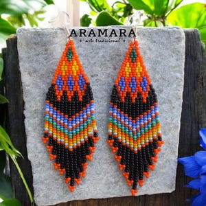 Puede incluir: Un par de pendientes de cuentas con forma de hoja. Los pendientes presentan un vibrante patrón de cuentas naranjas, negras, azules, amarillas y verdes. El texto "ARAMARA arte tradicional" es visible. Los pendientes cuelgan de ganchos plateados.