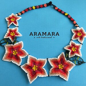 May include: A beaded necklace with a floral design. The necklace is made of white, red, orange, and yellow beads. The necklace is on a blue background. The text "ARAMARA arte tradicional" is visible in the center of the image.