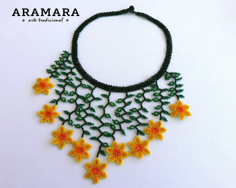 Op de afbeelding: Handgemaakte kralenketting met een zwarte kraag en een bloemmotief. De ketting heeft groene kralenranken en gele bloemen. De merknaam "ARAMARA" is linksboven zichtbaar. Een uniek sieraad.