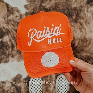 Puede incluir: Gorra de camionero naranja con texto bordado en blanco que dice "Raisin' Hell". La gorra tiene una parte trasera de malla blanca y una etiqueta blanca con el texto "The Bevel Label" y cinco estrellas.