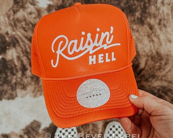 May include: Orange trucker hat with white embroidered text that reads "Raisin' Hell". The hat has a white mesh back and a white label with the text "The Bevel Label" and five stars.