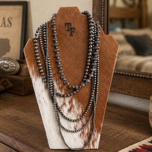 May include: A multi-strand necklace featuring dark metallic beads of varying sizes and textures, displayed on a brown and white cowhide-covered bust. The necklace consists of several layered strands of round and ribbed beads draped in a cascading fashion.