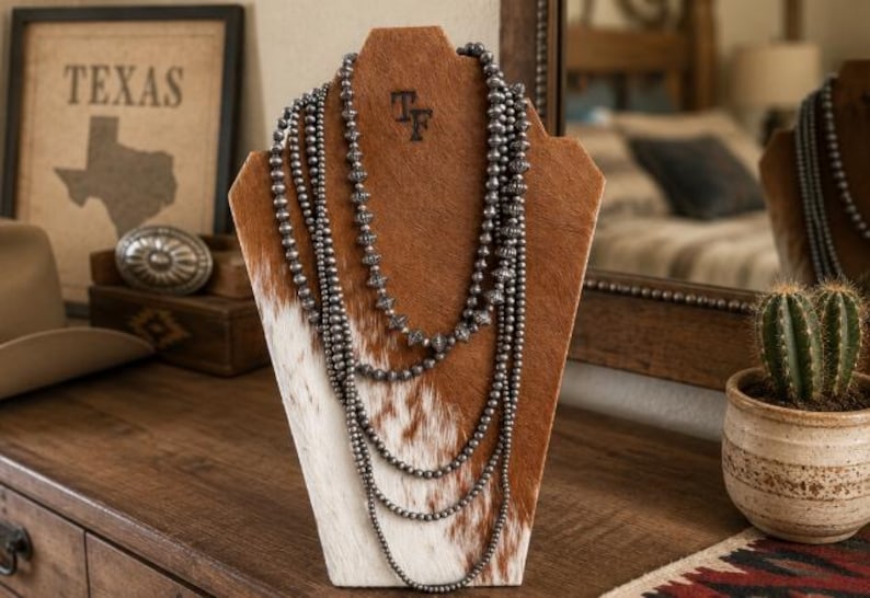 May include: A multi-strand necklace featuring dark metallic beads of varying sizes and textures, displayed on a brown and white cowhide-covered bust. The necklace consists of several layered strands of round and ribbed beads draped in a cascading fashion.