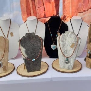 May include: A display of various necklaces on white and textured mannequin busts, including leather cords with metal pendants, turquoise accents, and heart-shaped charms, alongside a wooden rotating rack filled with hanging earrings on cards.