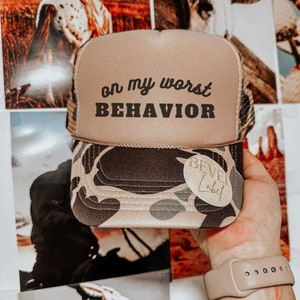 Può includere: Un cappello da camionista beige e mimetico con un logo ricamato nero che dice "Bevel Label" e il testo "on my worst BEHAVIOR" in nero.