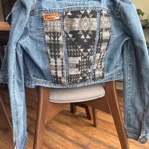 May include: A light blue denim jacket with a patterned back panel. The back panel features a geometric design in shades of black, gray, and brown. A small leather tag on the back reads "Artist Smith Designs". The jacket is displayed on a wooden chair.