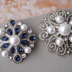 May include: Two silver brooches with pearl and crystal embellishments. One brooch features a snowflake design with blue crystals, while the other has a floral design with white pearls.