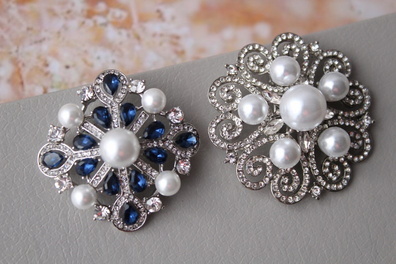 May include: Two silver brooches with pearl and crystal embellishments. One brooch features a snowflake design with blue crystals, while the other has a floral design with white pearls.