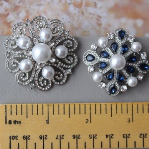 May include: Two silver brooches with pearl and rhinestone accents. The brooch on the left has a floral design with white pearls. The brooch on the right has a starburst design with blue rhinestones and white pearls. Both brooches are approximately 1 inch in diameter.