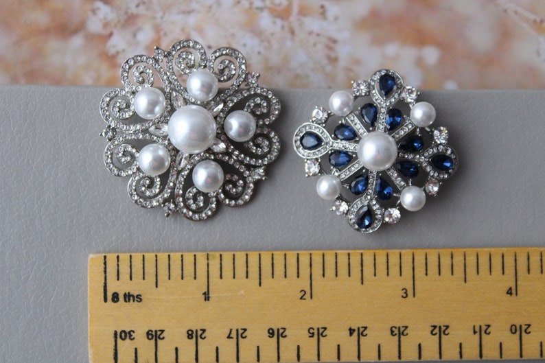 May include: Two silver brooches with pearl and rhinestone accents. The brooch on the left has a floral design with white pearls. The brooch on the right has a starburst design with blue rhinestones and white pearls. Both brooches are approximately 1 inch in diameter.