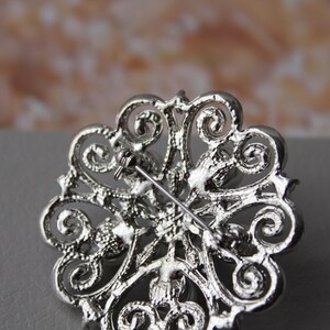 May include: Silver brooch with a filigree design and clear rhinestones. The brooch has a pin back.