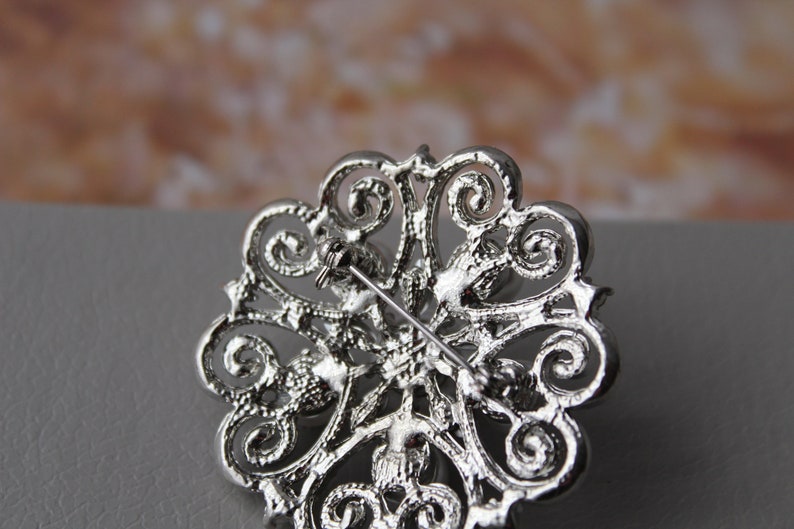 May include: Silver brooch with a filigree design and clear rhinestones. The brooch has a pin back.