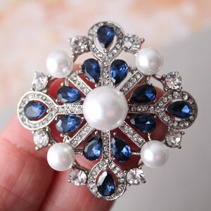 May include: A silver and blue brooch with a large pearl in the center. The brooch is decorated with smaller pearls and blue rhinestones.
