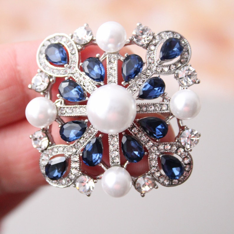 May include: A silver brooch with a square design featuring a large white pearl in the center surrounded by smaller blue and clear rhinestones. The brooch is decorated with a delicate filigree pattern.