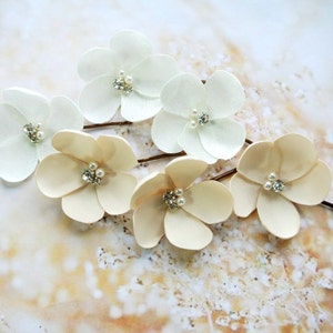 May include: Six white and ivory satin flower hair pins with pearl and rhinestone accents. The pins are arranged on a light brown background.