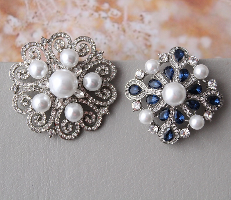 May include: Two silver brooches with pearl and crystal accents. One brooch features a floral design with white pearls, while the other has a snowflake design with blue crystals.