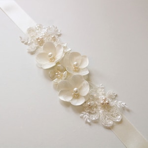 May include: A white satin ribbon with a floral embellishment. The embellishment features white fabric flowers with pearl accents and delicate lace.