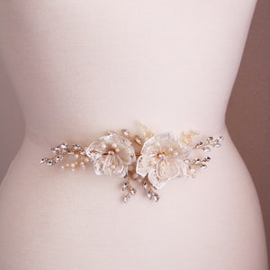 May include: A delicate bridal belt with a floral design featuring two white flowers with pearl and crystal embellishments. The belt is adorned with gold wire and crystal accents.