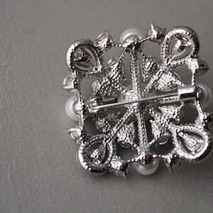 May include: A silver brooch with a floral design and white pearls. The brooch has a pin on the back for attaching to clothing.