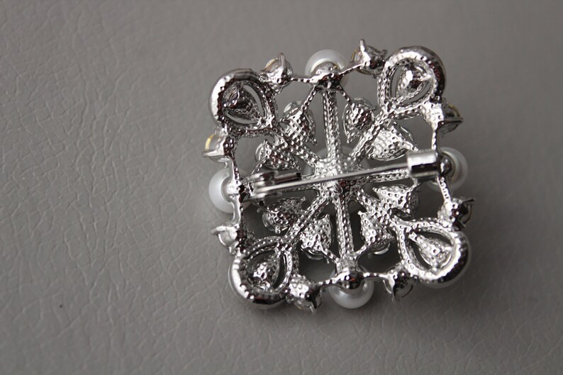 May include: A silver brooch with a floral design and white pearls. The brooch has a pin on the back for attaching to clothing.