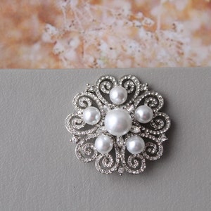 May include: A silver brooch with a floral design and white pearls. The brooch is on a gray background.