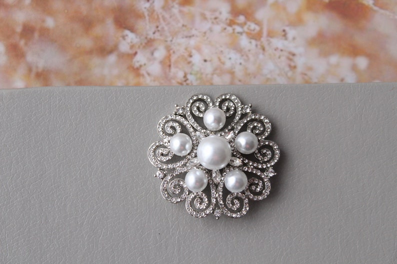 May include: A silver brooch with a floral design and white pearls. The brooch is on a gray background.