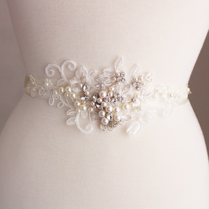 May include: A white satin bridal sash featuring a delicate lace design and pearl and rhinestone embellishments.