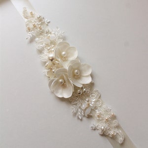 May include: A white satin ribbon with a delicate lace applique featuring white flowers and pearl accents.